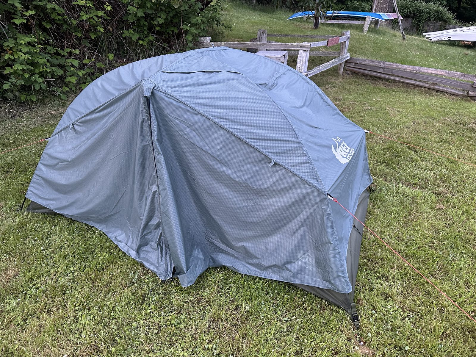 A grey REI Co-op tent pitched on grass, with paddleboards resting on a rack behind a wooden fence
