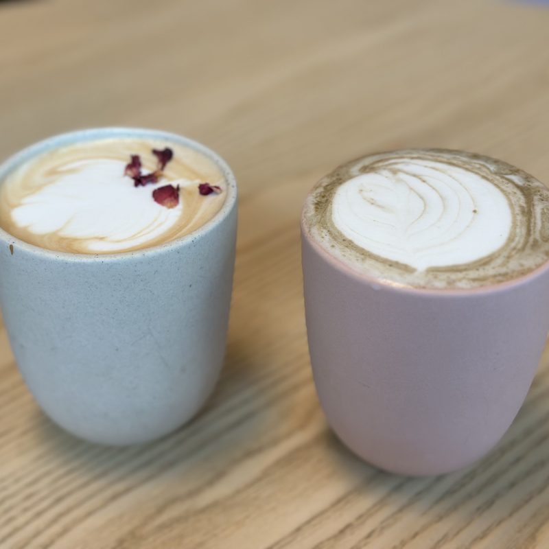 A blue latte and a pink hojicha side by side on pale wood