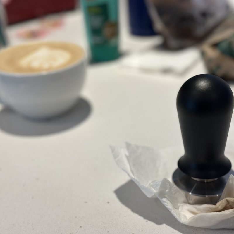 Espresso tamper standing on a white counter, a finished latte out of focus behind