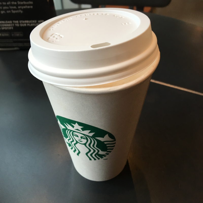 Lidded Starbucks paper cup on a black caf&eacute; table, in-store signage behind